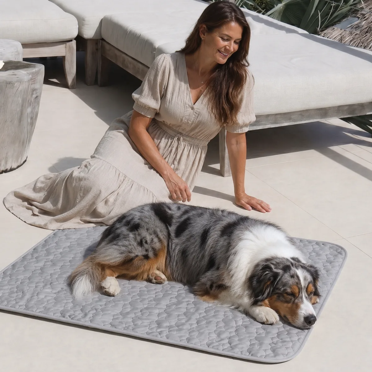 Berger australien endormi sur un tapis rafraîchissant gris posé sur la terrasse, sa propriétaire assise à côté de lui au soleil